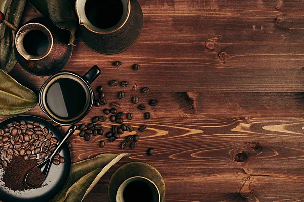 Café quente em xícara preta e vários potes turcos cezve com feijão, folhas secas com espaço de cópia no fundo de madeira velha placa marrom, vista superior . — Fotografia de Stock