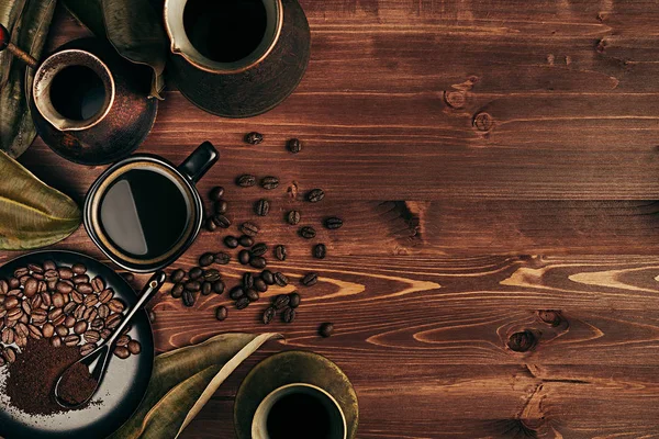 Hot coffee in black cup and several turkish pots cezve with beans, dry leaves with copy space on brown old wooden board background, top view. — Stock Photo, Image