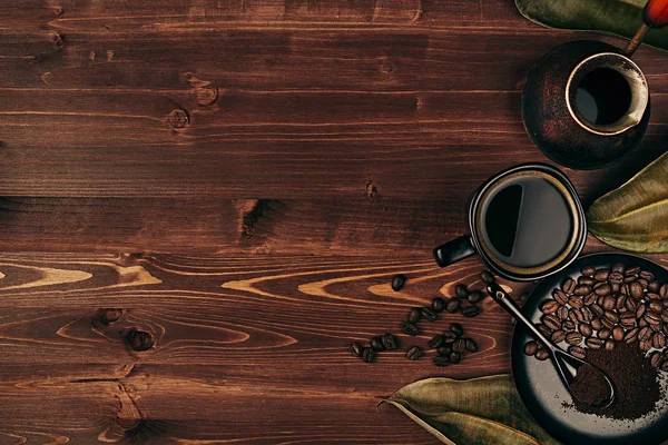 Горячий кофе в черной чашке с бобами, сухие листья и турецкий горшок cezve с копией пространства на коричневом старом деревянном фоне доски, вид сверху — стоковое фото