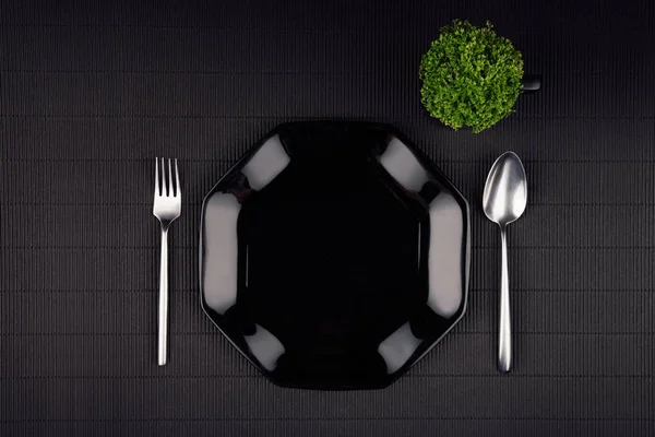 Sötét, modern, minimalista étterem menü modell, akár a fekete fényes lemez, kanalat, villát és zöld növény, felülnézet. — Stock Fotó