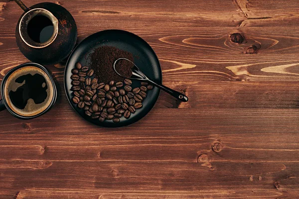 Hot coffee in black cup with beans, spoon and turkish pot cezve with copy space on brown old wooden board background, top view. — Stock Photo, Image
