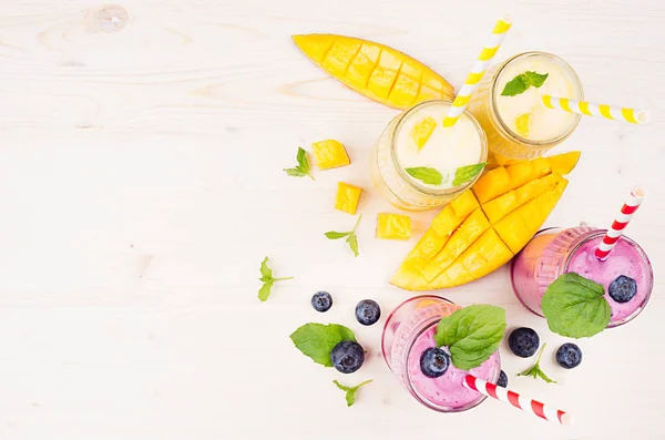 Свіжозмішаний жовтий і фіолетовий фруктовий смузі в скляних банках з соломою, листя м'яти, скибочки манго і ягоди, вид зверху . — стокове фото