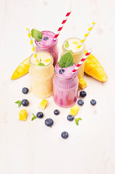 Batido de frutas amarillas y violetas recién mezclado en frascos de vidrio con paja, hojas de menta, rodajas de mango, arándanos. Fondo de tablero de madera blanco suave . —  Fotos de Stock