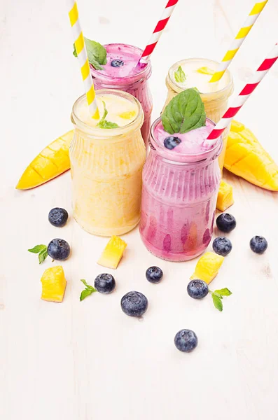 Batido de frutas amarillas y violetas en frascos de vidrio con paja, hojas de menta, rodajas de mango, bayas. Fondo de tablero de madera blanco suave . —  Fotos de Stock