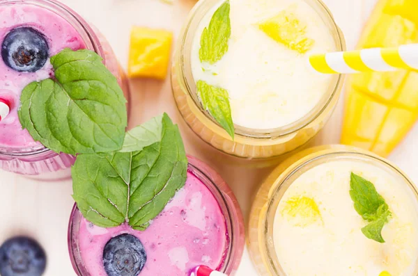 Batido de frutas amarillas y violetas recién mezclado en frascos de vidrio con paja, hojas de menta, rodajas de mango y bayas, de cerca, vista superior. Fondo de tablero de madera blanco suave . —  Fotos de Stock