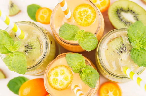 Čerstvě směsových oranžová kumquat a ovocné smoothie ve skleněných nádobách se slámou, list máty, zelené kiwi řez zralé bobule, pohled shora, zblízka. Dřevěná deska bílé barvy pozadí. — Stock fotografie