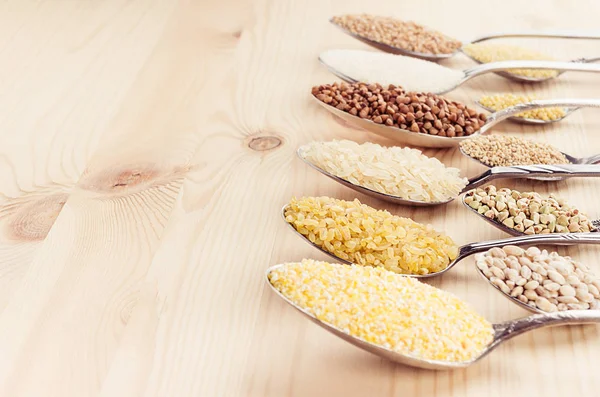 Grano de cereales diferentes en cucharas sobre tabla de madera beige, fondo rústico . — Foto de Stock