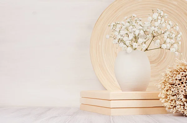 Soft home decor of beige bamboo dish, twigs and white small flowers in ceramic vase  on white wood background. I