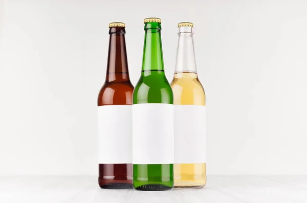 Bottiglie di birra 500ml di diversi colori con etichetta bianca bianca bianca su cartone di legno bianco, modellare. Modello per la pubblicità, il design, l'identità di branding . — Foto Stock