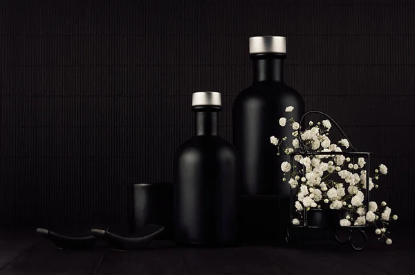 Dark black minimalist interior with blank black  bottles, white small flowers on dark wood board, mock up.