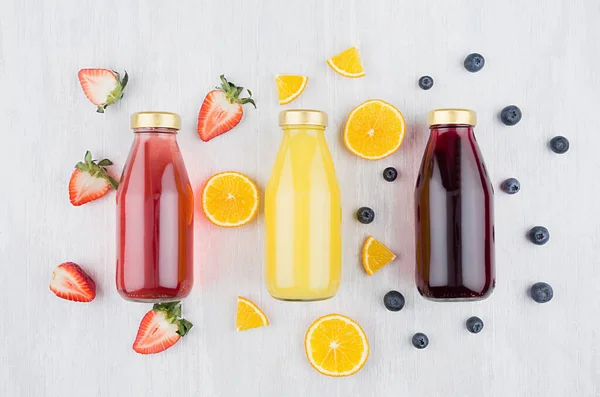 Organic yellow orange, pink strawberry and violet blueberry juices in glass bottles with fruit ingredients on white wood board, mock up for design, advertising.