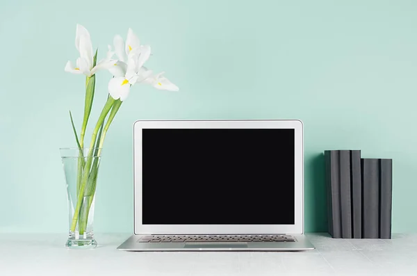 Spring home office for business with blank notebook monitor, black books and white fresh flowers in light green mint menthe interior on white wood desk.