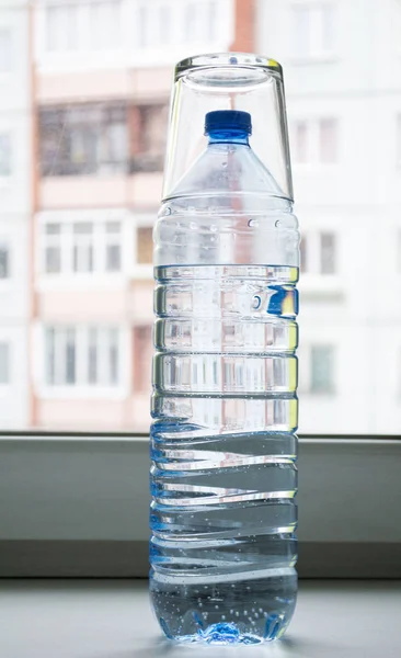 plastic bottle with water on top covered with a glass cup on a white windowsill