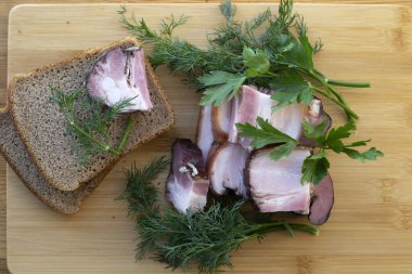 Tahta bir tahtanın üzerinde tütsülenmiş et, ekmek, dereotlu ve maydanozlu. Pencere önünde pastırmalı pano. en üstteki görünüm