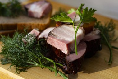 Tahta bir tahtanın üzerinde tütsülenmiş et, ekmek, dereotlu ve maydanozlu. Pencere önünde pastırmalı pano. güneşli bir gün.