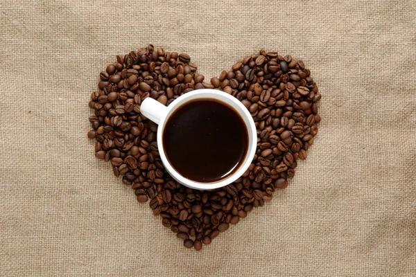 heart made from coffee beans top in the center a white cup with coffee. sackcloth background. view from above