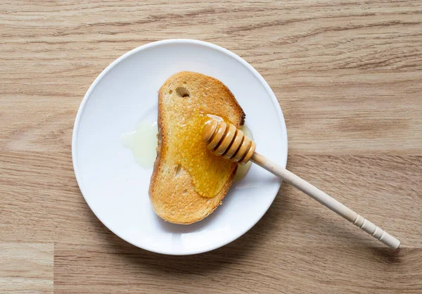 Vara de madeira com mel em uma torradeira e um pires branco. mel pingando de um pau de mel em uma torradeira . — Fotografia de Stock