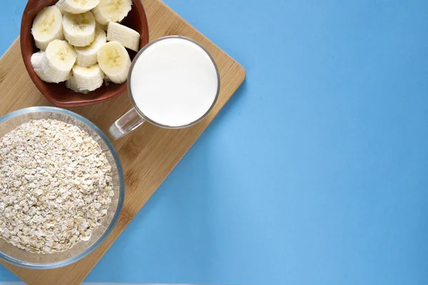 sliced banana slices in a clay plate, a glass mug with milk, a glass bowl with oatmeal on a wooden board and a blue background. Healthy breakfast. the right food. flat lay copy space.