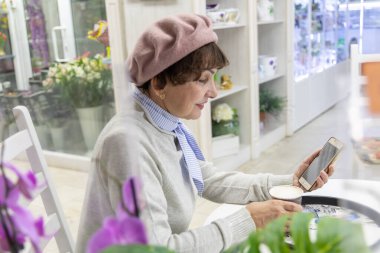 Bereli, mutlu, beyaz, yaşlı bir kadın, gülümsüyor, kafede bir masada otururken cep telefonuyla konuşuyor. Ön planda orkide çiçekleri var. Yan görünüm