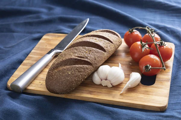 Fresh bread, tomato, garlic, knife on a cutting board. Dark blue background. Side view. Closeup
