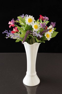 Beyaz vazodaki yapay çiçekler. Siyah arka planda beyaz bir vazoda suni çiçek buketi..