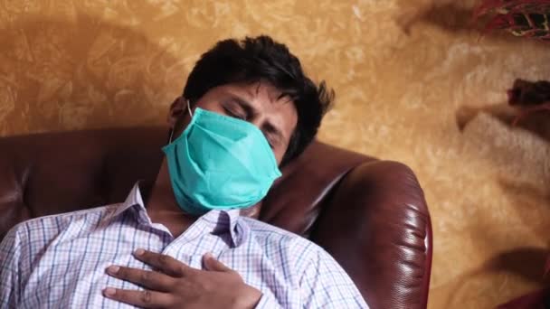 Больной человек с головной болью в защитной маске сидит на диване — стоковое видео