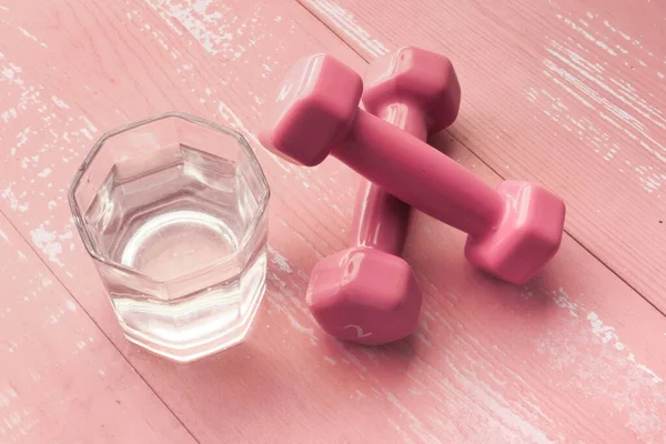 Zbliżenie różowy kolor hantel i szklanka wody na stole — Zdjęcie stockowe