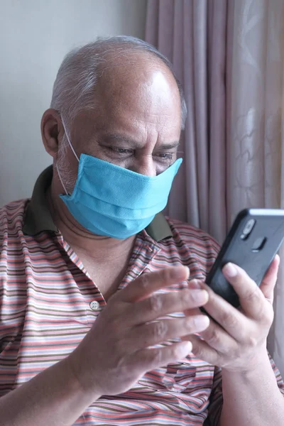 Ανώτερος άνθρωπος με μάσκα προσώπου χρησιμοποιώντας έξυπνο τηλέφωνο στο σπίτι — Φωτογραφία Αρχείου