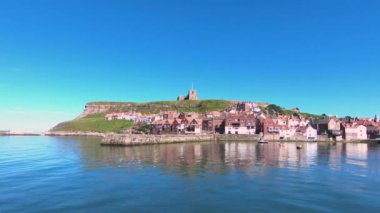 Whitby Rıhtımı. Whitby rıhtımının karşısındaki manzara, Kuzey İngiltere 'de bir Kuzey Yorkshire sahil kasabası. St. Mary Kilisesi, Doğu Kayalıkları 'nın zirvesinde görülebilir..