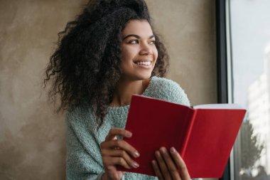 Başarılı bir üniversite öğrencisi, öğrenim dili, kütüphanede oturuyor. Güzel Afro-Amerikan kadın portresi kırmızı kitap tutuyor, pencereye bakıyor ve gülümsüyor. Sınav hazırlama konsepti