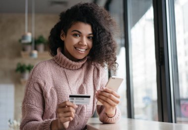 Güzel Afrikalı Amerikalı kadın kredi kartı tutuyor, ödeme yapıyor, kameraya bakıyor ve gülümsüyor. Mobil bankacılık konsepti. Genç mutlu kız akıllı telefon kullanıyor, internetten alışveriş yapıyor, bilet alıyor.
