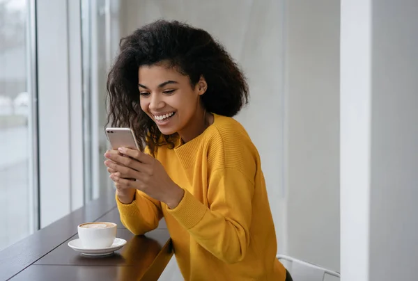 Beautiful African American girl with happy emotional face using smartphone, reading text message, sitting in modern cafe. Stylish positive woman holding mobile phone, shopping online, booking tickets