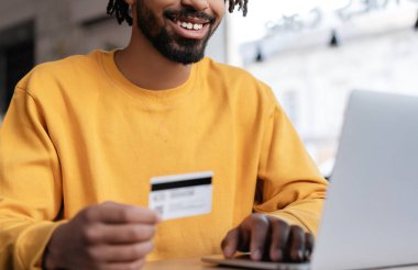 Happy African American man holding credit card, using laptop computer for online shopping. Hipster guy ordering food online, booking tickets and smiling. Successful freelancer receive payment 