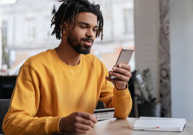 Kredi kartı tutan Afrikalı Amerikalı bir adam online alışveriş ve mobil bankacılık için akıllı telefon kullanıyor. Genç serbest çalışan ödeme alıyor, bakiyeyi kontrol ediyor. Düşünceli hipster internetten yemek sipariş ediyor, bilet ayırtıyor.