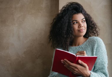 Güzel Afro-Amerikan kadın elinde kırmızı kitapla pencereye bakıyor ve gülümsüyor. Üniversite öğrencisiyim, dil öğreniyorum, kütüphanede oturuyorum. Not alan genç düşünceli yazarın portresi
