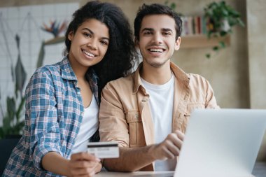 İnternetten alışveriş yapan, dizüstü bilgisayar kullanan, kameraya bakıp gülümseyen çok ırklı güzel bir çiftin portresi. Mutlu arkadaşlar internet sitesinden yemek sipariş ediyor, bilet ayırtıyor, kredi kartı tutuyor.