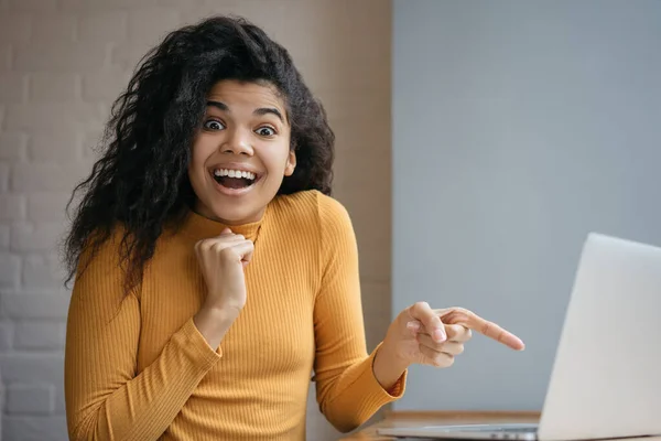 Surprised African American woman showing finger on laptop screen. Portrait of excited successful blogger influencer looking at camera with open mouth and happy emotional face
