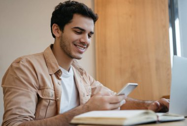 Hintli serbest çalışan, elinde akıllı telefon, iletişim, evden çalışıyor. Laptop, internet, planlama projesi kullanan yakışıklı Asyalı adam modern ofiste oturuyor. 