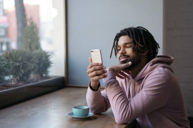 Genç yakışıklı Afrikalı Amerikalı adam cep telefonu kullanıyor, video izliyor, iletişim kuruyor, kafede oturuyor. Evde başarılı bir serbest çalışan.