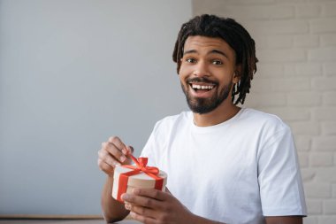 Genç yakışıklı Afrikalı Amerikalı adam doğum günü hediyesi tutuyor ve gülümsüyor, evde oturuyor. Mutlu duygusal adamın portresi hediye kutusunu açıyor.