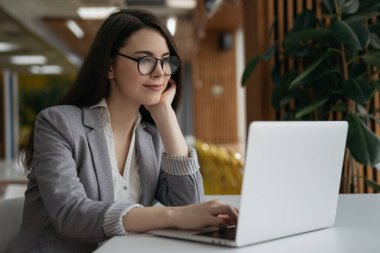 Düşünceli iş kadını şık gözlükler takıyor dizüstü bilgisayar kullanıyor, plan yapıyor, internette çalışıyor. İş yerindeki başarılı serbest çalışan beyin fırtınasının portresi