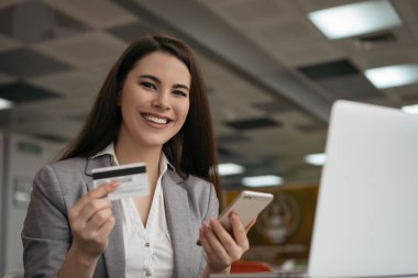 Kredi kartı tutan bir iş kadını, online alışveriş için akıllı telefonu kullanıyor, ofisinde oturuyor. Serbest çalışan ödeme alır, bakiyeyi kontrol eder. Gülümseyen kadın yemek ısmarlıyor, internet sitesinden bilet alıyor.