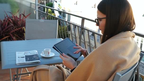 Portrait of young caucasian woman covered in blanket using ipad tablet app, drinking coffee,checking email online at terrace. slow motion. France, Nice. Woman at self isolation quarantine reading last news