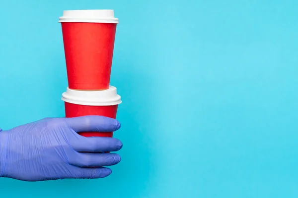 Hand of delivery man medical gloves carrying paper cups coffee for takeaway isolated on blue background. contactless delivery service in quarantine coronavirus pandemic. courier volunteer donation.