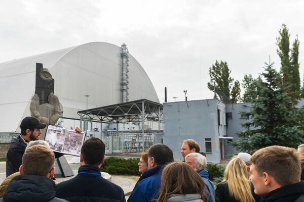 Мужской гид перед Чернобыльским мемориалом показывает фотографию блока ядерных реакторов после взрыва. Чернобыльская АЭС стоит на заднем плане. Чернобыль.10.2019
