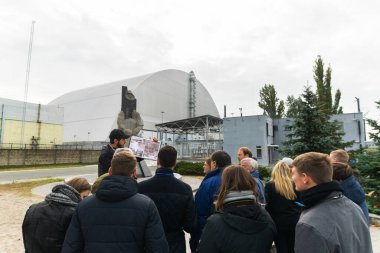 Group of tourists with a tour guide stands by the Block 4 reactor in Chernobyl.  Male person shows real photos after accident. Ukraine.08.10.2019