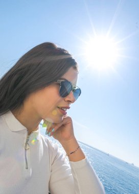 Portrait of a young girl with white shirt and sunglasses who is holding her hand on her cheek and admiring the vies. Background with sunflare and the sea.