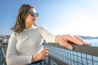 Sexy brunette girl is posing with her hands on the fence and turned  towards the sea. Glamorous fashionable trendy background image.
