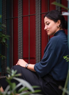 Young girl in elegant outfit sits on the stairs and poses in front of the red door and green wall and looks down calmly. Low angle photograph of A pretty middle eastern woman from Oman.