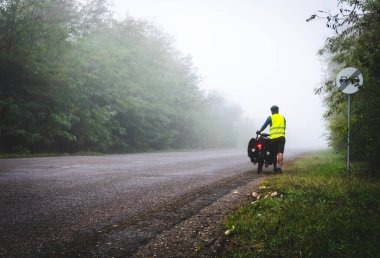 Cyclist with reflective vest and lighs on is walking down the road to the fog in nature. Bicycle touring in bad  weather conditions and bad visability.